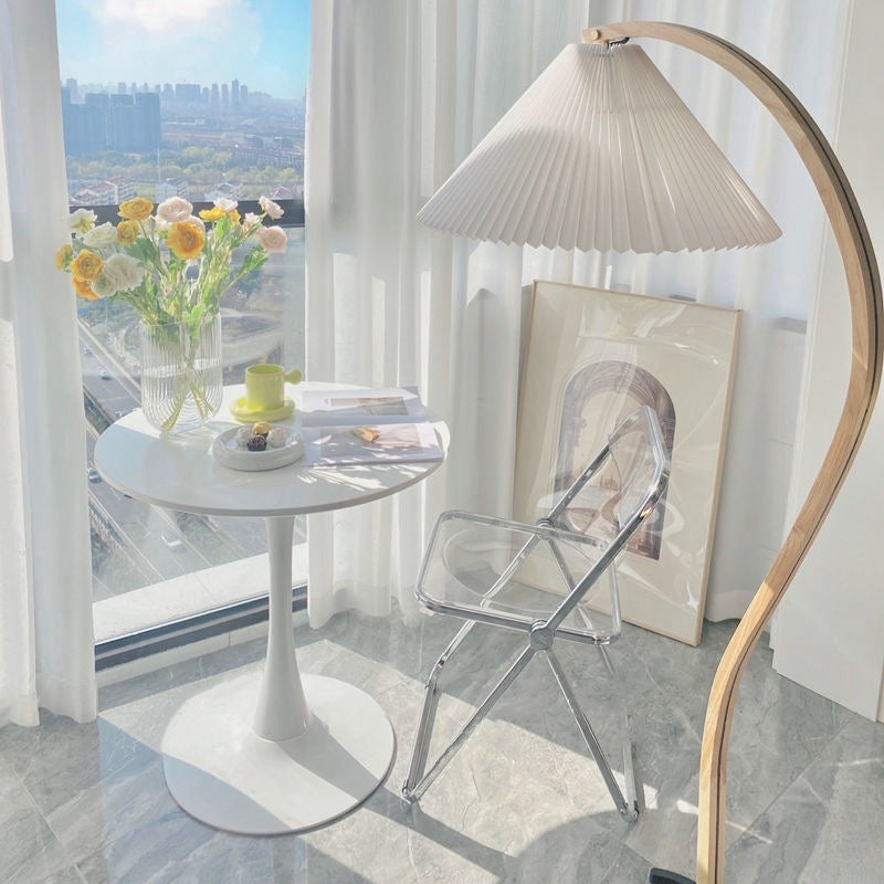 Small round dining or coffee table setup by a window with a transparent folding chair, a leather chair, and a floor lamp. Ideal for home or commercial environments like cafes or lounges. Suitable for wholesale furniture purchases directly from China.